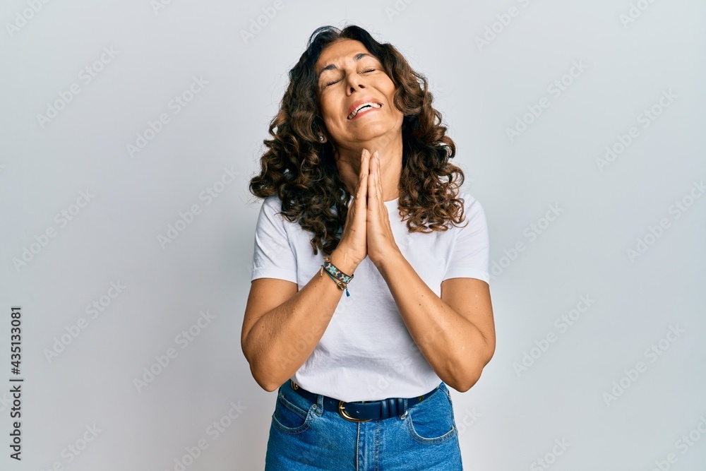 Middle age hispanic woman wearing casual white t shirt begging and praying with hands together with hope expression on face very emotional and worried. begging. Middle age hispanic woman wearing casual white t shirt begging and praying with hands together with hope expression on face very emotional and worried. begging.