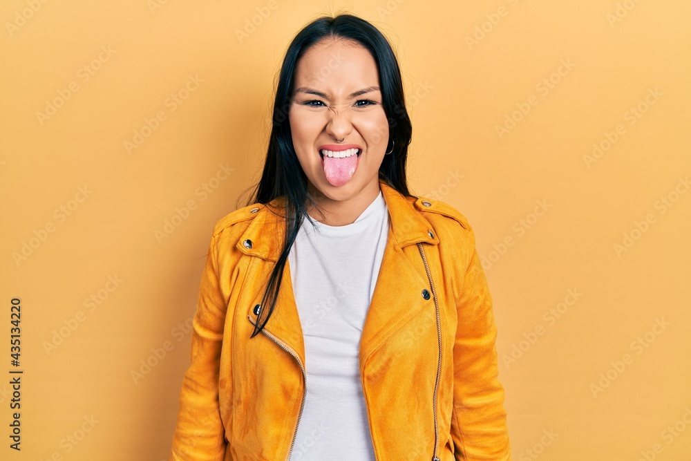 Beautiful hispanic woman with nose piercing wearing yellow leather jacket sticking tongue out happy with funny expression. emotion concept. Beautiful hispanic woman with nose piercing wearing yellow leather jacket sticking tongue out happy with funny expression. emotion concept.