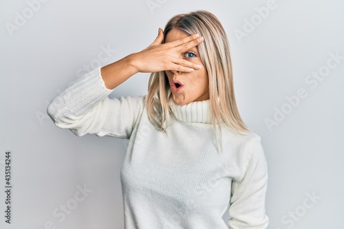 Beautiful blonde woman wearing casual turtleneck sweater peeking in shock covering face and eyes with hand, looking through fingers afraid