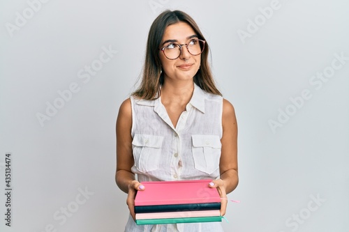 Young hispanic woman holding books smiling looking to the side and staring away thinking.