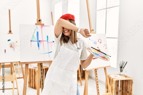 Young artist woman painting on a canvas at art studio smiling cheerful playing peek a boo with hands showing face. surprised and exited photo