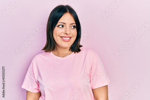 Young hispanic woman wearing casual pink t shirt looking away to side with smile on face, natural expression. laughing confident.