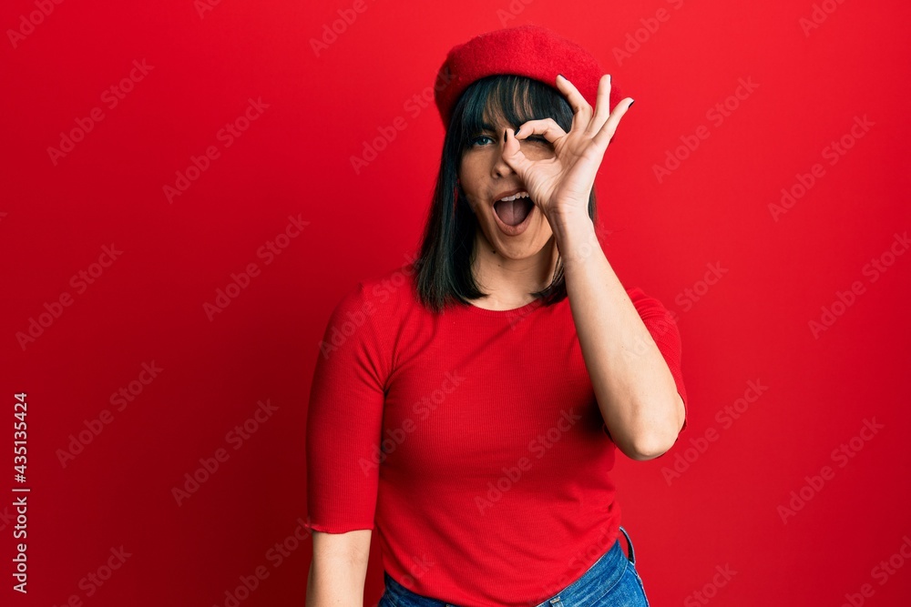 Young hispanic woman wearing french look with beret doing ok gesture with hand smiling, eye looking through fingers with happy face. Young hispanic woman wearing french look with beret doing ok gesture with hand smiling, eye looking through fingers with happy face.