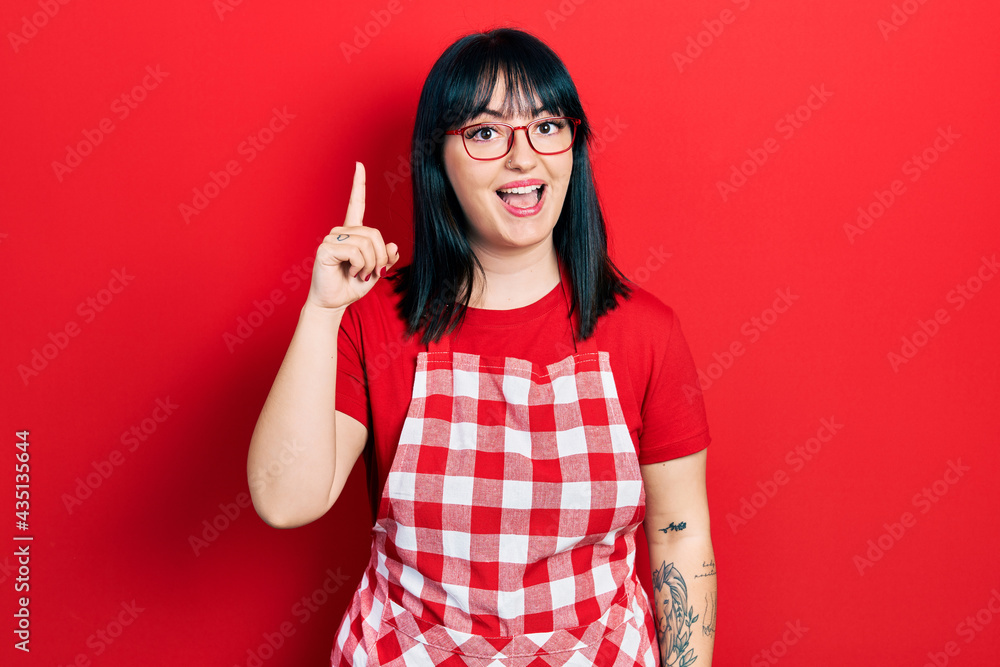 Young hispanic woman wearing cook apron and glasses pointing finger up with successful idea. exited and happy. number one. Young hispanic woman wearing cook apron and glasses pointing finger up with successful idea. exited and happy. number one.