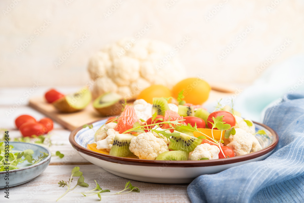 Vegetarian salad of cauliflower cabbage, kiwi, tomatoes, microgreen sprouts on gray wooden background. Side view, selective focus.