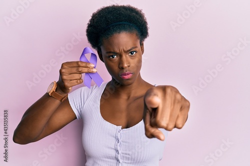 Young african american girl holding purple ribbon awareness pointing with finger to the camera and to you, confident gesture looking serious © Krakenimages.com