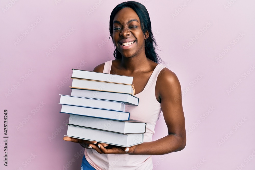 Young african american woman holding books winking looking at the camera with sexy expression, cheerful and happy face. Young african american woman holding books winking looking at the camera with sexy expression, cheerful and happy face.