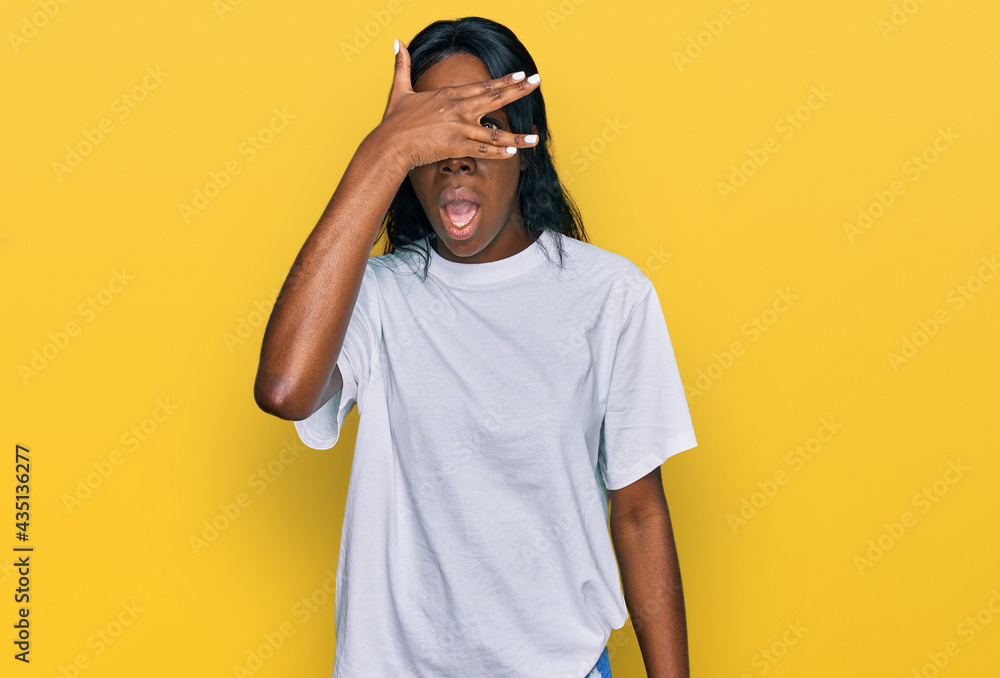 Fototapeta premium Young african american woman wearing casual white t shirt peeking in shock covering face and eyes with hand, looking through fingers with embarrassed expression.