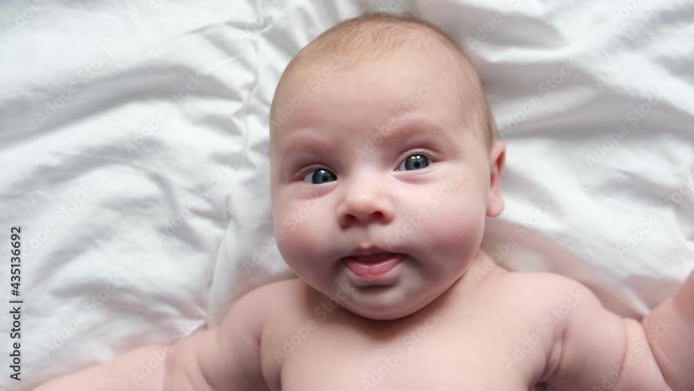 Close-up portrait a newborn girl. The face of Caucasian infant baby lying on the bed, showing his tongue, and waving his arms. One month baby looking at camera. Little child relaxing. Head portrait. Close-up portrait a newborn girl. The face of Caucasian infant baby lying on the bed, showing his tongue, and waving his arms. One month baby looking at camera. Little child relaxing. Head portrait.