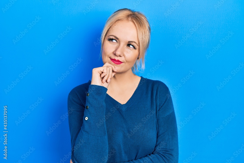 Young blonde woman wearing casual blue sweater thinking concentrated about doubt with finger on chin and looking up wondering Young blonde woman wearing casual blue sweater thinking concentrated about doubt with finger on chin and looking up wondering