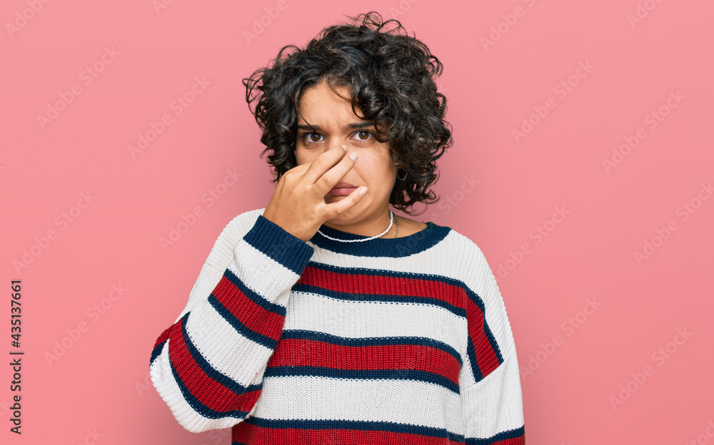 Young hispanic woman with curly hair wearing casual clothes smelling something stinky and disgusting, intolerable smell, holding breath with fingers on nose. bad smell Young hispanic woman with curly hair wearing casual clothes smelling something stinky and disgusting, intolerable smell, holding breath with fingers on nose. bad smell
