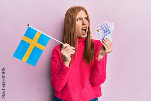 Young irish woman holding sweden flag and krone banknotes angry and mad screaming frustrated and furious, shouting with anger. rage and aggressive concept.