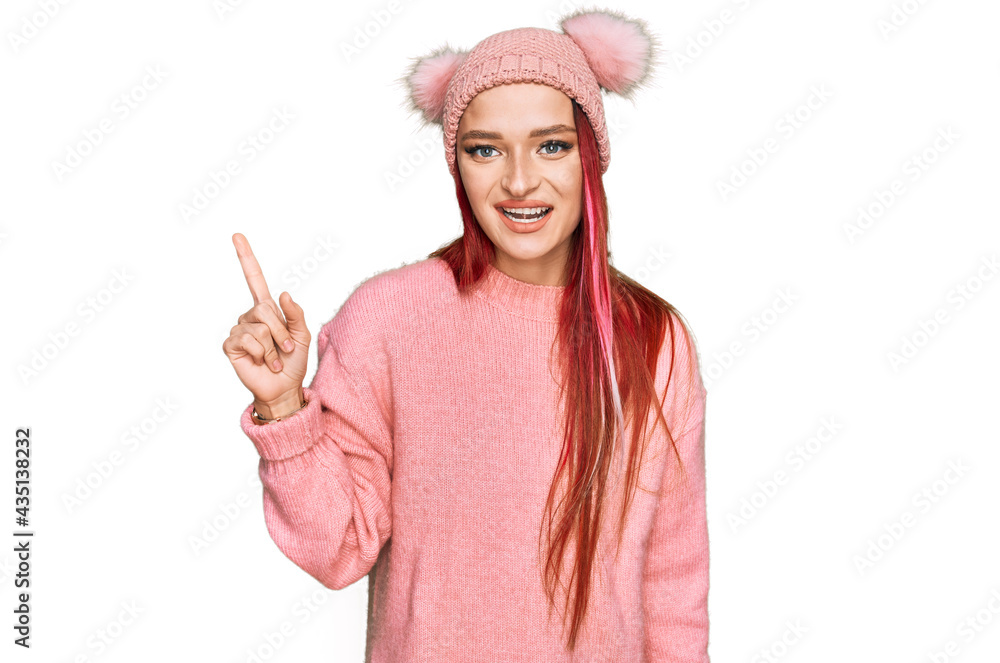 Young caucasian woman wearing casual clothes and wool cap pointing finger up with successful idea. exited and happy. number one. Young caucasian woman wearing casual clothes and wool cap pointing finger up with successful idea. exited and happy. number one.