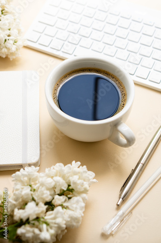 Beige and white work space with coffee cup, notepad and lilac flowers