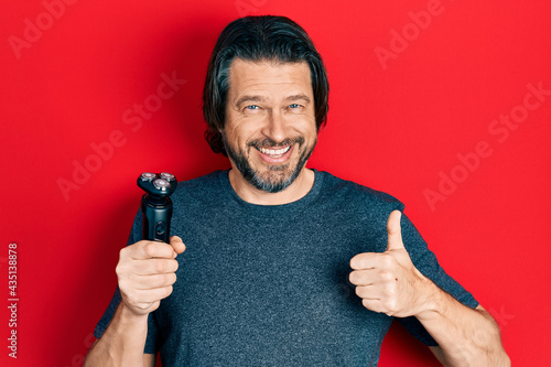 Middle age caucasian man holding electric razor machine smiling happy and positive, thumb up doing excellent and approval sign