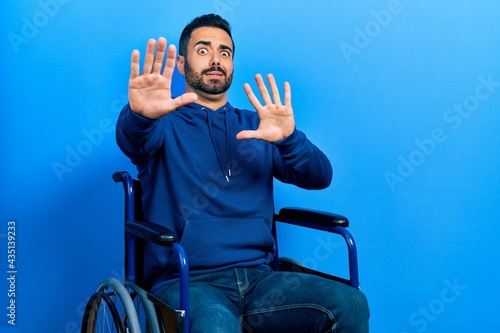 Handsome hispanic man with beard sitting on wheelchair afraid and terrified with fear expression stop gesture with hands, shouting in shock. panic concept.