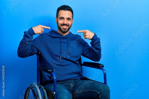 Handsome hispanic man with beard sitting on wheelchair smiling cheerful showing and pointing with fingers teeth and mouth. dental health concept.