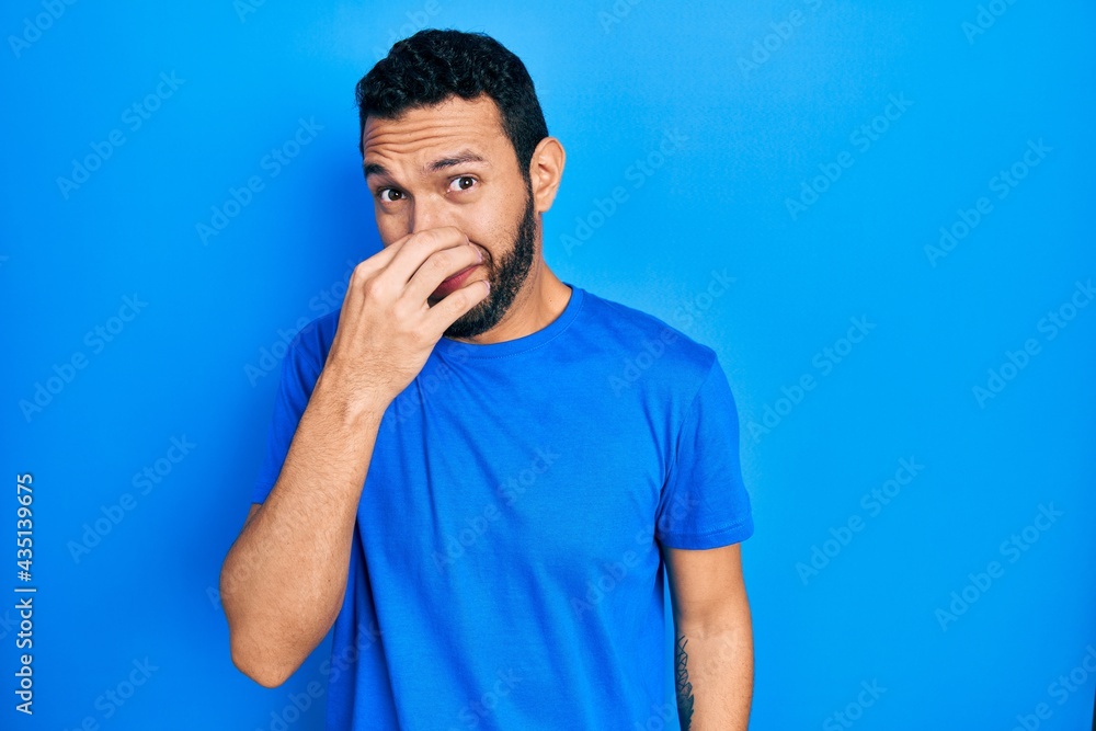 Hispanic man with beard wearing casual blue t shirt smelling something stinky and disgusting, intolerable smell, holding breath with fingers on nose. bad smell Hispanic man with beard wearing casual blue t shirt smelling something stinky and disgusting, intolerable smell, holding breath with fingers on nose. bad smell