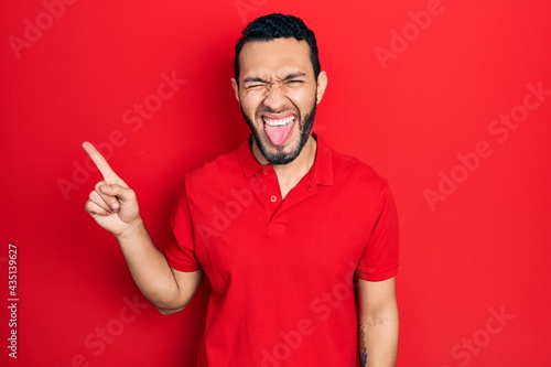 Hispanic man with beard pointing with fingers to the side sticking tongue out happy with funny expression.