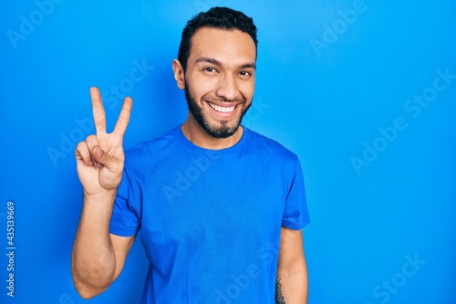 Hispanic man with beard wearing casual blue t shirt showing and pointing up with fingers number two while smiling confident and happy.