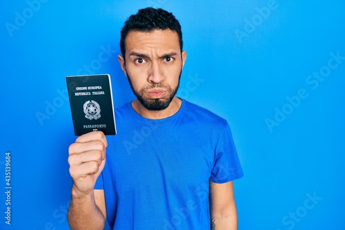Hispanic man with beard holding italy passport puffing cheeks with funny face. mouth inflated with air, crazy expression.