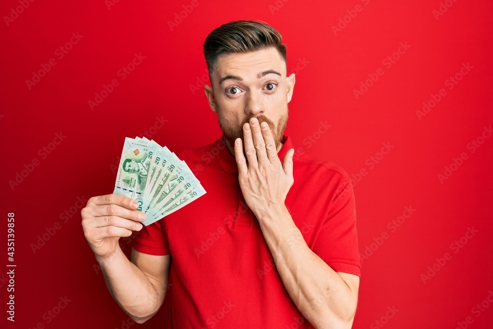 Young redhead man holding 20 thai baht banknotes covering mouth with hand, shocked and afraid for mistake. surprised expression Young redhead man holding 20 thai baht banknotes covering mouth with hand, shocked and afraid for mistake. surprised expression