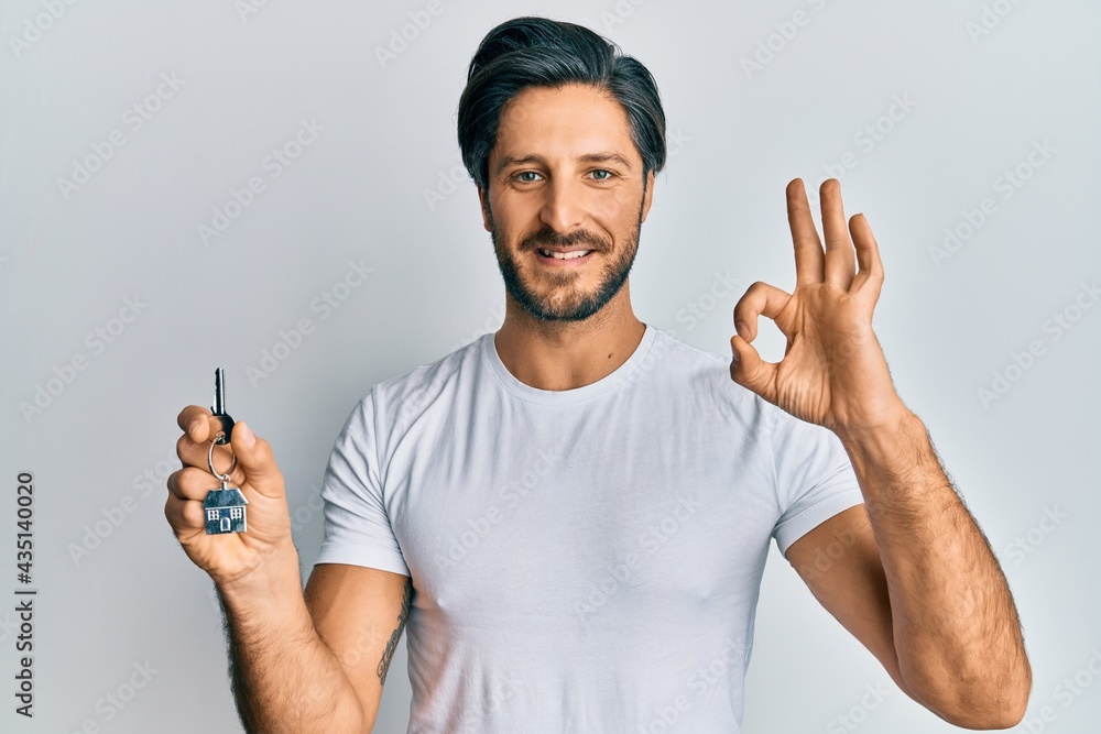Young hispanic man holding keys of new home doing ok sign with fingers, smiling friendly gesturing excellent symbol Young hispanic man holding keys of new home doing ok sign with fingers, smiling friendly gesturing excellent symbol