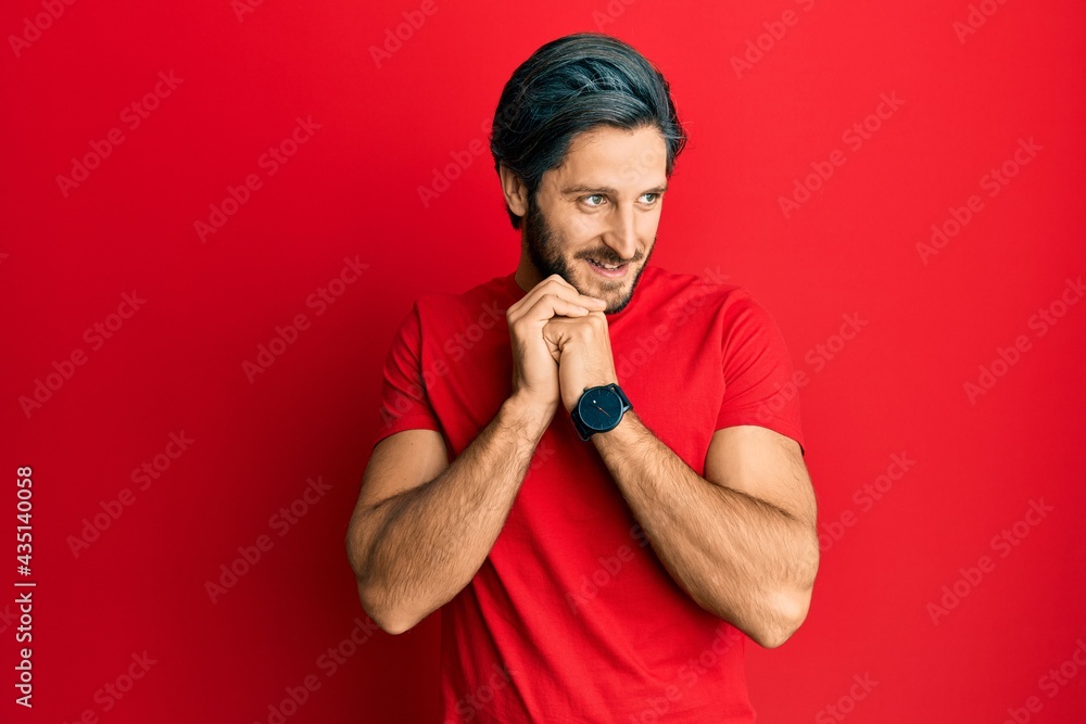 Young hispanic man wearing casual red t shirt laughing nervous and excited with hands on chin looking to the side
