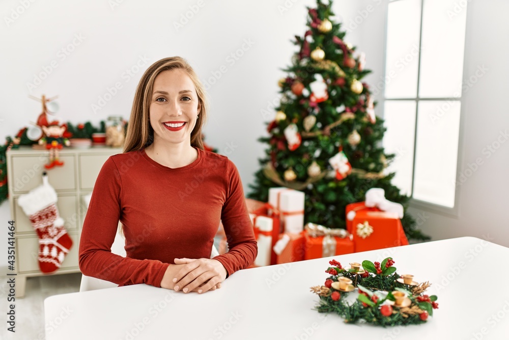 Caucasian young blonde woman sitting on the table by christmas tree with a happy and cool smile on face. lucky person. Caucasian young blonde woman sitting on the table by christmas tree with a happy and cool smile on face. lucky person.