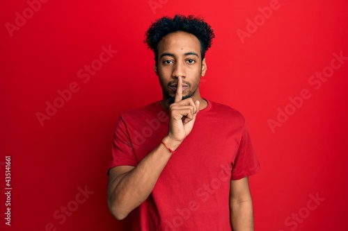 Young african american man with beard wearing casual red t shirt asking to be quiet with finger on lips. silence and secret concept.