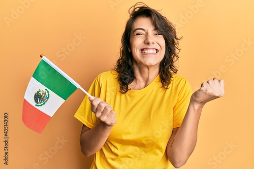 Young hispanic woman holding mexico flag screaming proud  celebrating victory and success very excited with raised arm