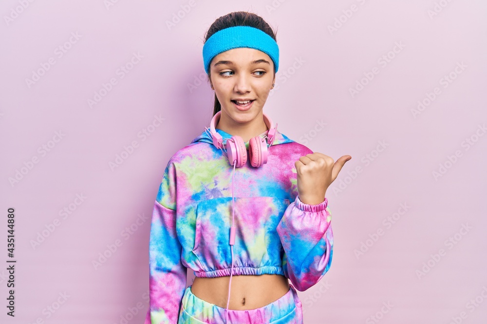 Young brunette girl wearing gym clothes and headphones smiling with happy face looking and pointing to the side with thumb up. Young brunette girl wearing gym clothes and headphones smiling with happy face looking and pointing to the side with thumb up.