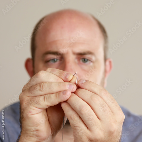 Close-up shot of man's hands threading a sewing needle with a thread for sewing clothes. Selective focus on hands. Male person doing sewing. Household chores.