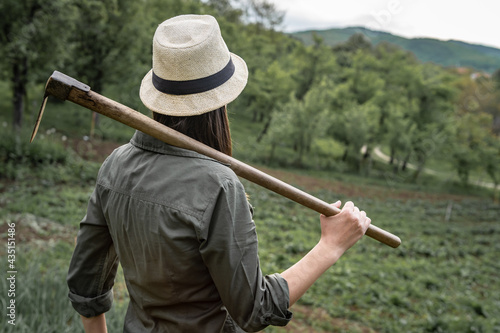Back view portrait of young adult caucasian woman female farmer standing in the agricultural field preparing plants posing with hoe on her shoulder in summer day looking to the side with copy space