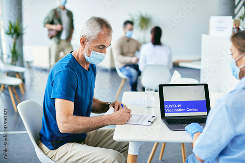 Mature man filling medical record during COVID-19 vaccination procedure at vaccination center. © Drazen
