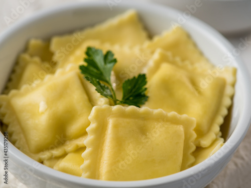 The photo shows ravioli in a white earthenware plate. Above is a green sprig of parsley. Close-up. Macro photography. There are no people in the photo. Bright lighting.