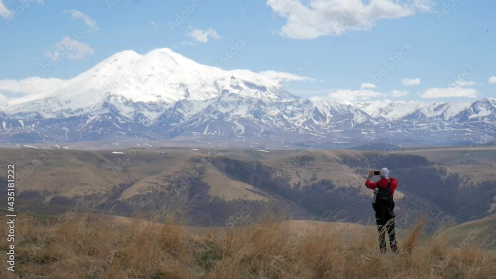 a male tourist traveler on a hill overlooking the mountain range of the Caucasus mountains Elbrus stands boldly on a rock and takes pictures on his phone a male tourist traveler on a hill overlooking the mountain range of the Caucasus mountains Elbrus stands boldly on a rock and takes pictures on his phone