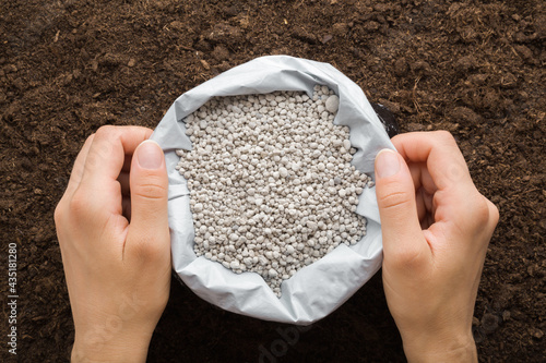 Young adult woman hands holding opened plastic bag with gray complex fertiliser granules on dark soil background. Closeup. Product for root feeding of vegetables, flowers and plants. Top down view. photo