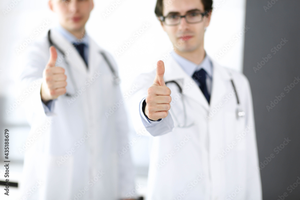 Two male doctors standing as a team and showing thumbs up as a symbol of the best service for patients in the clinic. Medicine and health care Two male doctors standing as a team and showing thumbs up as a symbol of the best service for patients in the clinic. Medicine and health care