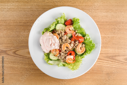 Salad with shrimps, quinoa, tomatoes, peppers, cucumber, lettuce, mayonnaise on white round plate on wooden table