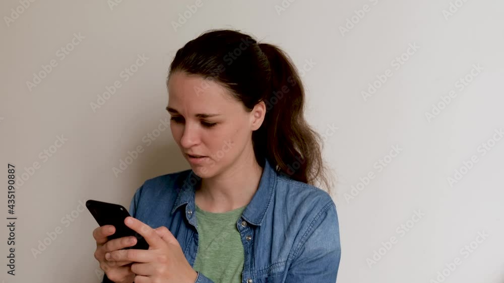 Young Caucasian European woman in shirt on white background is texting using smartphone and looking closely at phone screen. Important conversation and correspondence over Internet online. 4K footage. Young Caucasian European woman in shirt on white background is texting using smartphone and looking closely at phone screen. Important conversation and correspondence over Internet online. 4K footage.