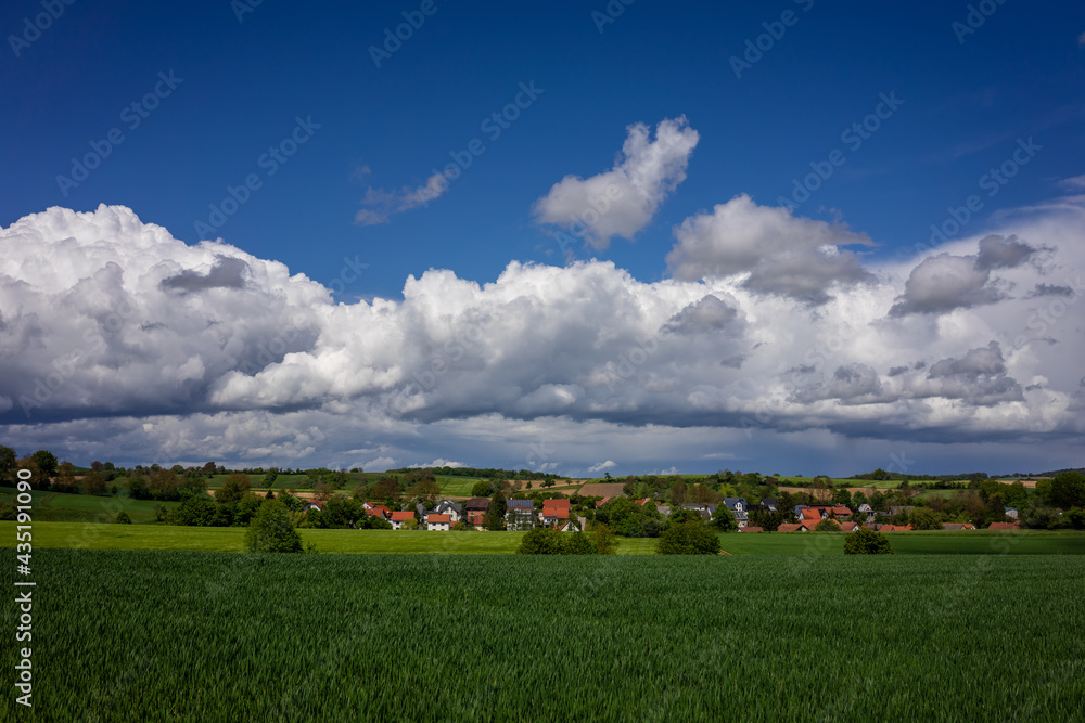 Obraz premium Schöne, grüne Frühlingslandschaft im Kraichgau, Deutschland, mit Getreidefeld im Vordergrund und einem Dorf mit bewölktem Himmel im Hintergrund.