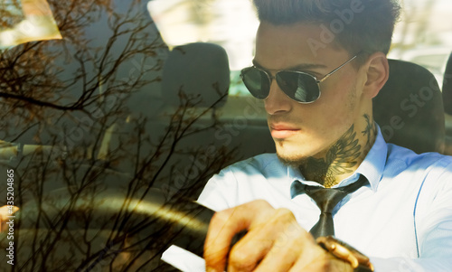 Young man with tattoo and sunglasses  in blue shirt sitting in car and holding steering wheel.