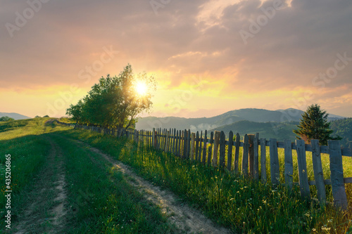 Beautiful summer evening scenery with a rural road on a hill and wooden fence along with it at green Carpathian mountains. Sun on a beautiful sunset sky is shining through the green trees.