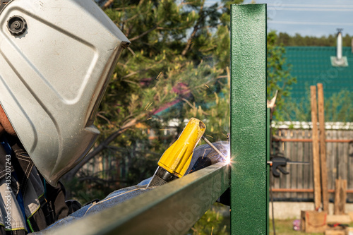 The welder welds a new metal frame for the fence around the house in backdrop pines trees, village photo