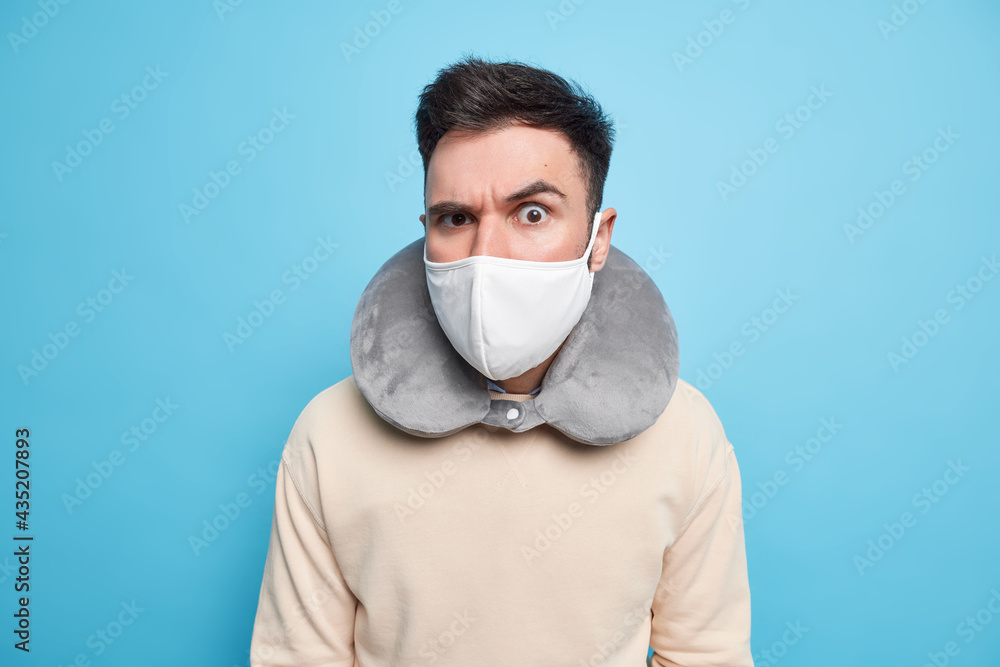 Indoor shot of serious attentive man wears protective mask against coronavirus inflated neck pillow for comfort sleep during travelling in transport raises eyebrow looks attentively at camera Indoor shot of serious attentive man wears protective mask against coronavirus inflated neck pillow for comfort sleep during travelling in transport raises eyebrow looks attentively at camera