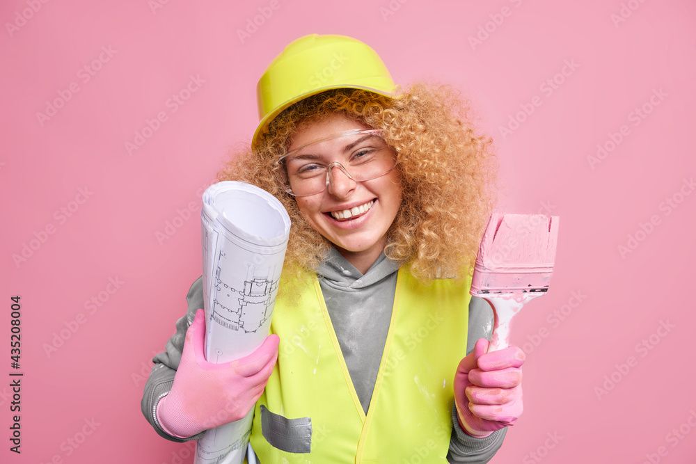 Cheerful construction worker being in good mood tries to implement her arhitectural plan in life holds blueprint and brush for painting walls wears protective helmet and uniform isolated on pink Cheerful construction worker being in good mood tries to implement her arhitectural plan in life holds blueprint and brush for painting walls wears protective helmet and uniform isolated on pink