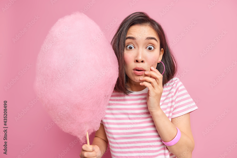 Stunned scared brunette woman holds sweet candy floss surprised to hear something amazing finds out how much calories she is going to eat wears striped t shirt isolated over pink background. Stunned scared brunette woman holds sweet candy floss surprised to hear something amazing finds out how much calories she is going to eat wears striped t shirt isolated over pink background.