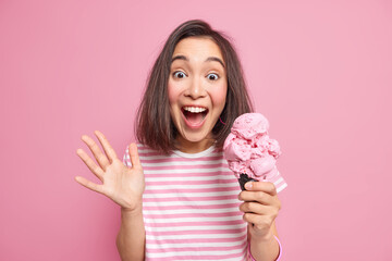 Emotional brunette woman exlaims loudly keeps palm raised eats delicious ice cream reacts on something unexpected has surpised joyfull look wears casual striped t shirt isolated over pink background