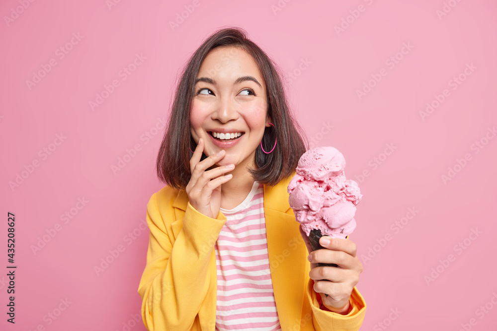 Horizontal shot of cheerful brunette young Asian woman looks away gladfully holds delicious cold ice cream in waffle wears elegant clothes enjoys summer holidays isolated over pink background. Horizontal shot of cheerful brunette young Asian woman looks away gladfully holds delicious cold ice cream in waffle wears elegant clothes enjoys summer holidays isolated over pink background.